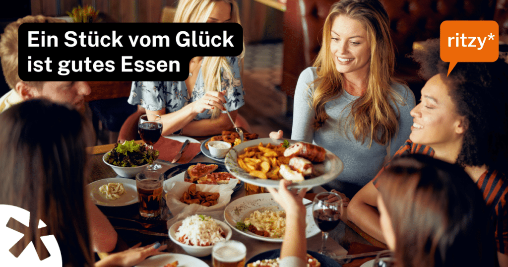 Gruppe von Personen sitzt gemeinsam an einem Tisch in einem Restaurant und teilt verschiedene Gerichte; darüber steht der Text „Ein Stück vom Glück ist gutes Essen“ sowie das ritzy*-Logo.